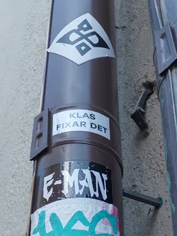 A sticker with an abstract geometric pattern in black and white on a diamond shape.  There are other stickers present on the same surface, including one that reads "KLAS FIXAR DET" and another with "E-MAN" written on it.