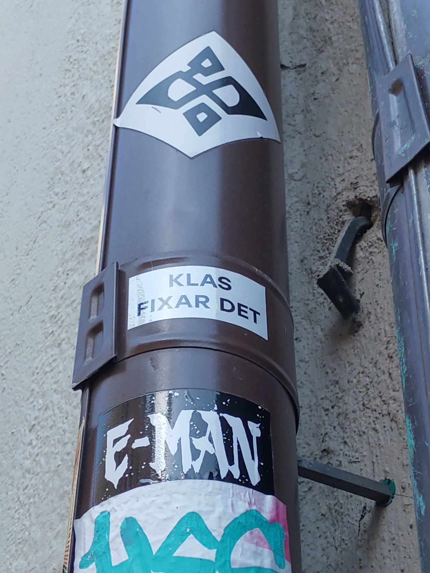 A sticker with an abstract geometric pattern in black and white on a diamond shape.  There are other stickers present on the same surface, including one that reads "KLAS FIXAR DET" and another with "E-MAN" written on it.