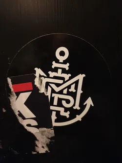 A black and white circular sticker featuring a unique anchor design incorporating stylized lettering. The anchor is detailed with decorative elements and the letters are intricately designed within the anchor form.  Part of another sticker is visible underneath.