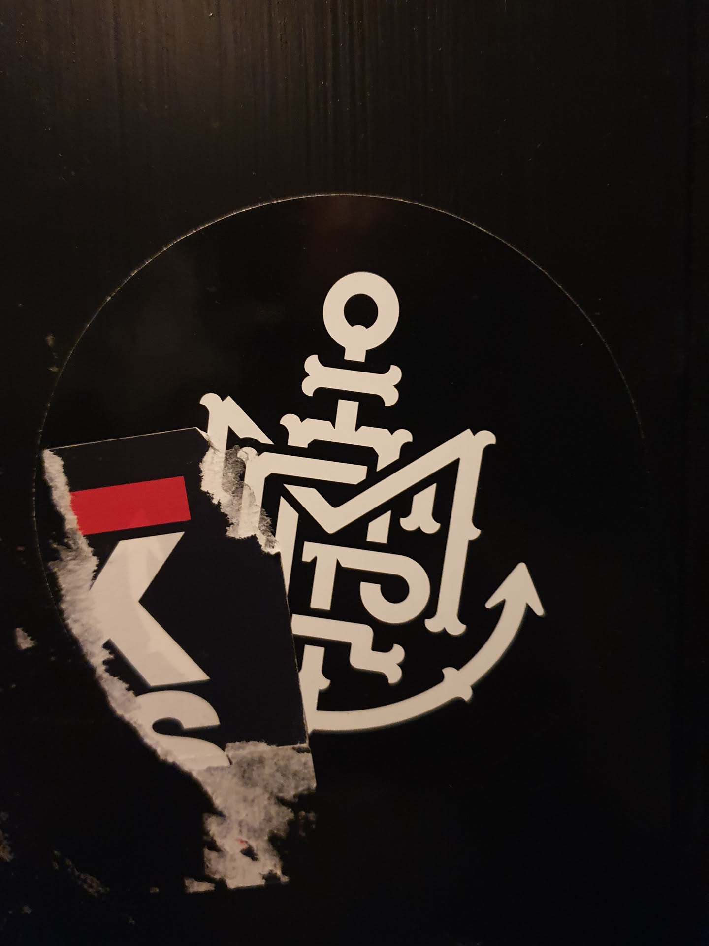 A black and white circular sticker featuring a unique anchor design incorporating stylized lettering. The anchor is detailed with decorative elements and the letters are intricately designed within the anchor form.  Part of another sticker is visible underneath.