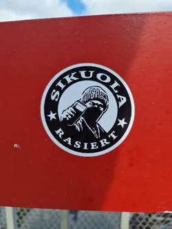 Close-up of a round sticker featuring a black and white illustration of a person in a beanie holding what appears to be a razor or similar tool. The words "SIKUOJA" and "RASIERT" are inscribed around the image in a circular fashion. The sticker is affixed to a red surface.