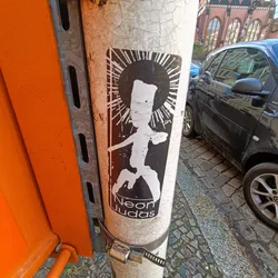 Black and white sticker with a figure reminiscent of a sun god or religious icon. The sticker is attached to a weathered pole.