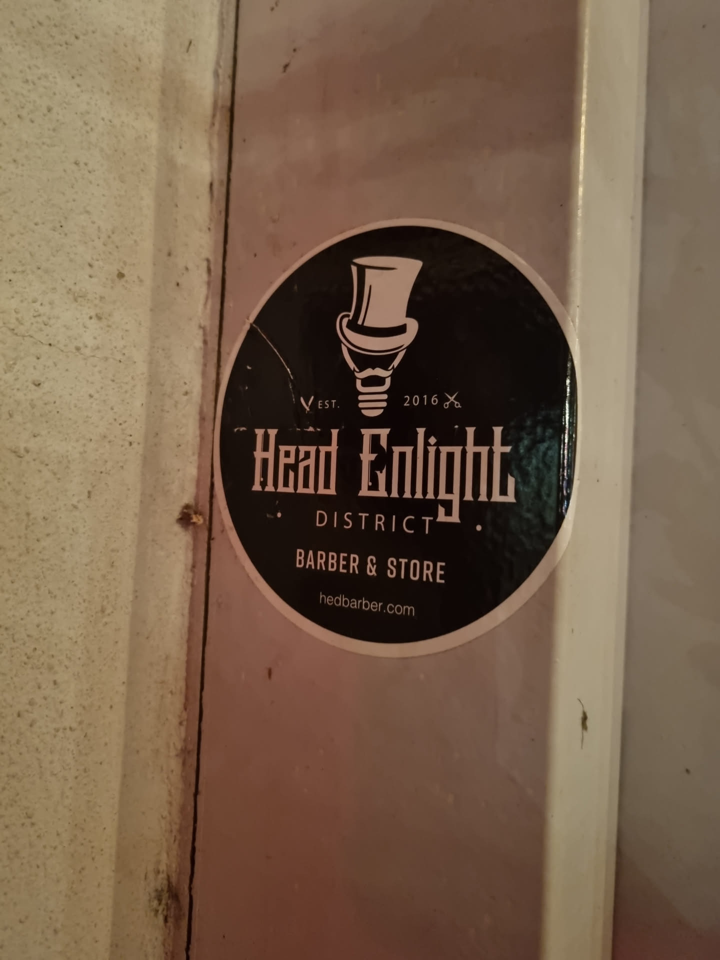 Round sticker with a black background and white design.  It features an illustration of a lightbulb in a top hat, the text "Headlight District", "Barber & Store", and the website address. 