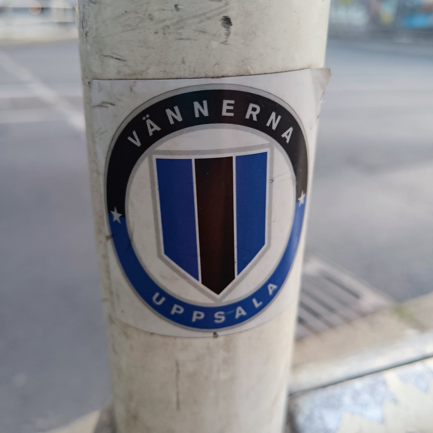 A round sticker with a central shield-shaped logo. The shield is primarily blue with a vertical black stripe.  The words "VÄNNERNA" and "UPPSALA" encircle the shield in a circular black design.  The sticker appears to be affixed to a pole outdoors.