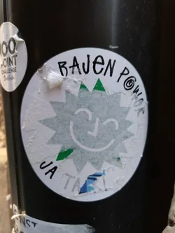 A round sticker with a smiling sun graphic and the text "BAJEN POWER" at the top.  There's also some partially visible text at the bottom that looks like "JA T...". The style is simple and playful.