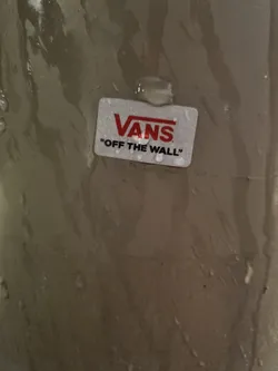 A small, rectangular Vans sticker with the Vans logo and the text "Off The Wall" is adhered to a surface. The surface appears to be wet or damp.