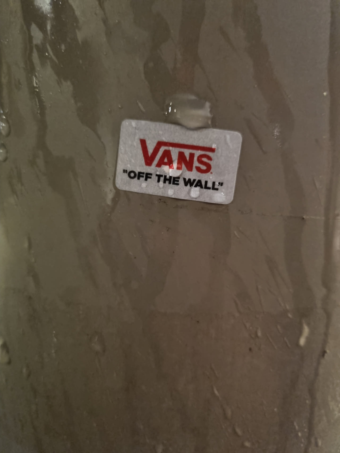 A small, rectangular Vans sticker with the Vans logo and the text "Off The Wall" is adhered to a surface. The surface appears to be wet or damp.