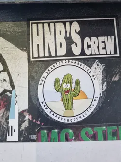 A round sticker featuring a cartoon cactus smiling in a desert landscape. The text on the sticker reads 'www.smartshopmarties.nl'.  There is also a separate sticker that says 'HNB'S Crew'.