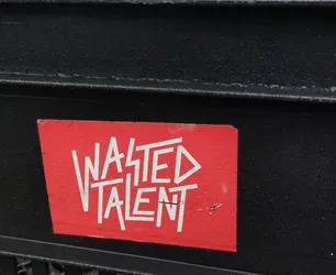 A red rectangular sticker with the text "WASTED TALENT" written in a stylized, bold white font. The sticker appears to be affixed to a dark-colored metal surface.