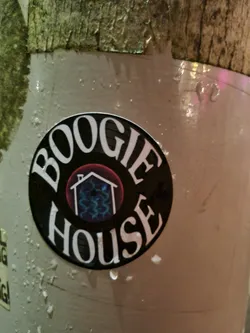 Round black and white sticker with the text "BOOGIE HOUSE" in a circular design.  It features a smaller graphic of a house with a red roof and blue wavy lines inside. The sticker appears to be affixed to a weathered surface.