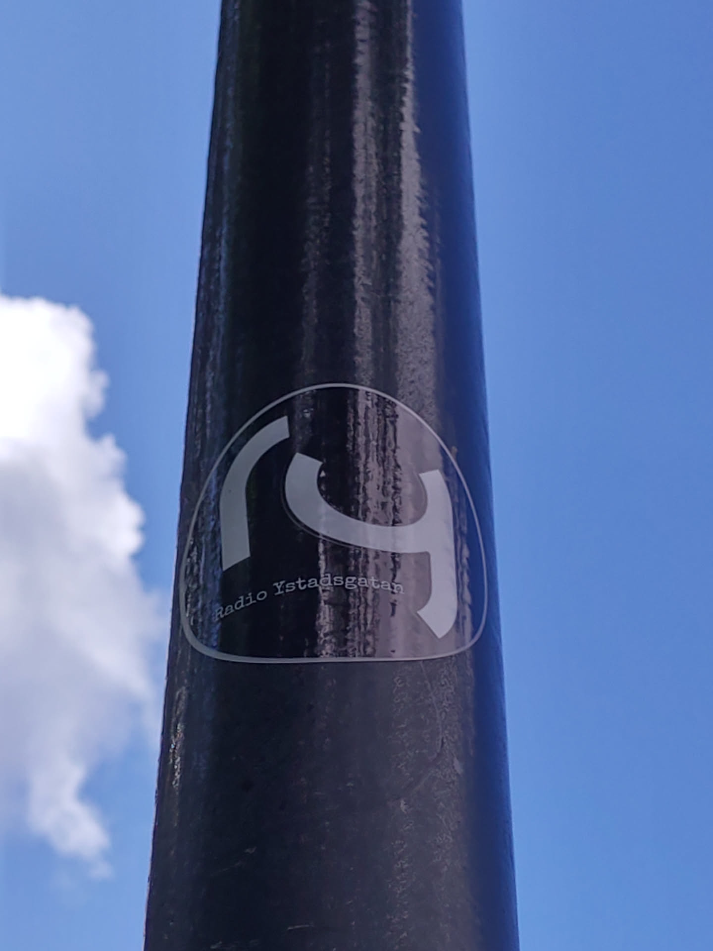 A black and white sticker with a logo and text on a dark pole. The sky is visible in the background.