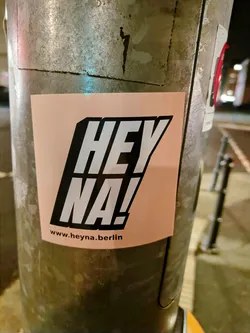 A peach-colored sticker with black and white text saying "HEY NA!" and the website www.heyona.berlin at the bottom. It is pasted on a metallic pole.