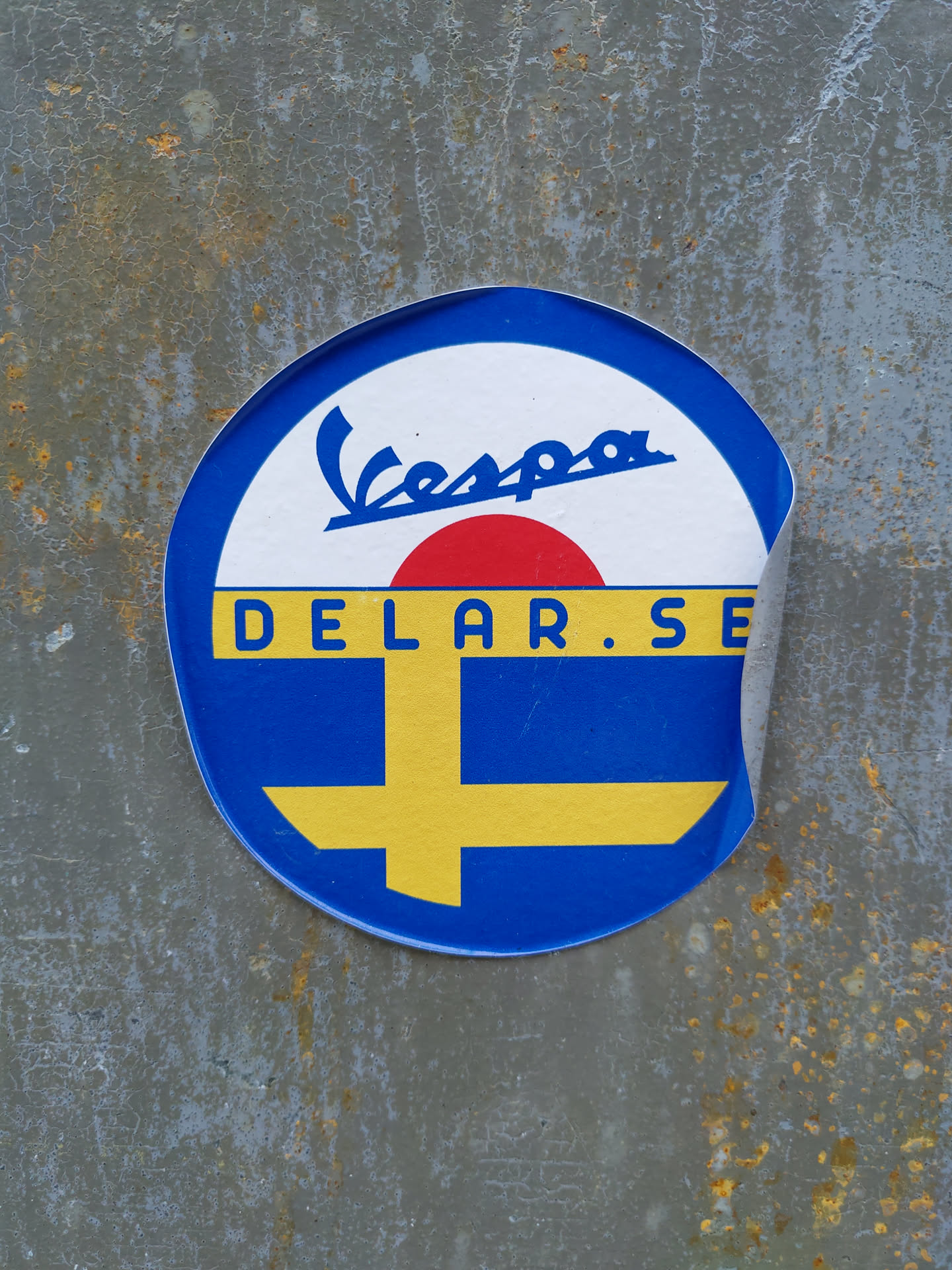 A round sticker featuring the Vespa logo and the text "Delar.se" is affixed to a rusty gray metal surface. The sticker's design incorporates blue, yellow, red, and white colors, with the yellow and blue resembling the Swedish flag. The sticker shows some signs of peeling at the edge.
