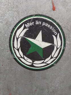 A round sticker with a green star in the center, surrounded by a white laurel wreath. The text "Mer än passion" is written around the star.