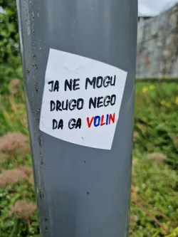 A sticker on a grey pole with a handwritten message in Serbian or Croatian. The text translates to "I can't do anything but love him".