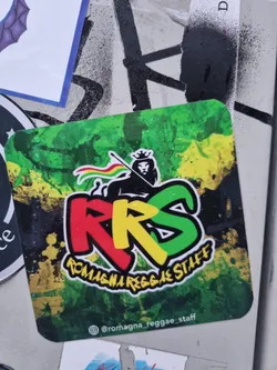 This square sticker features a vibrant design with a lion, the letters RRS, and the words "Romagna Reggae Staff" written in a stylized font. The colors are predominantly green, yellow, and red, reminiscent of the Jamaican flag. The background has a paint-splattered effect, and the overall style is bold and eye-catching.