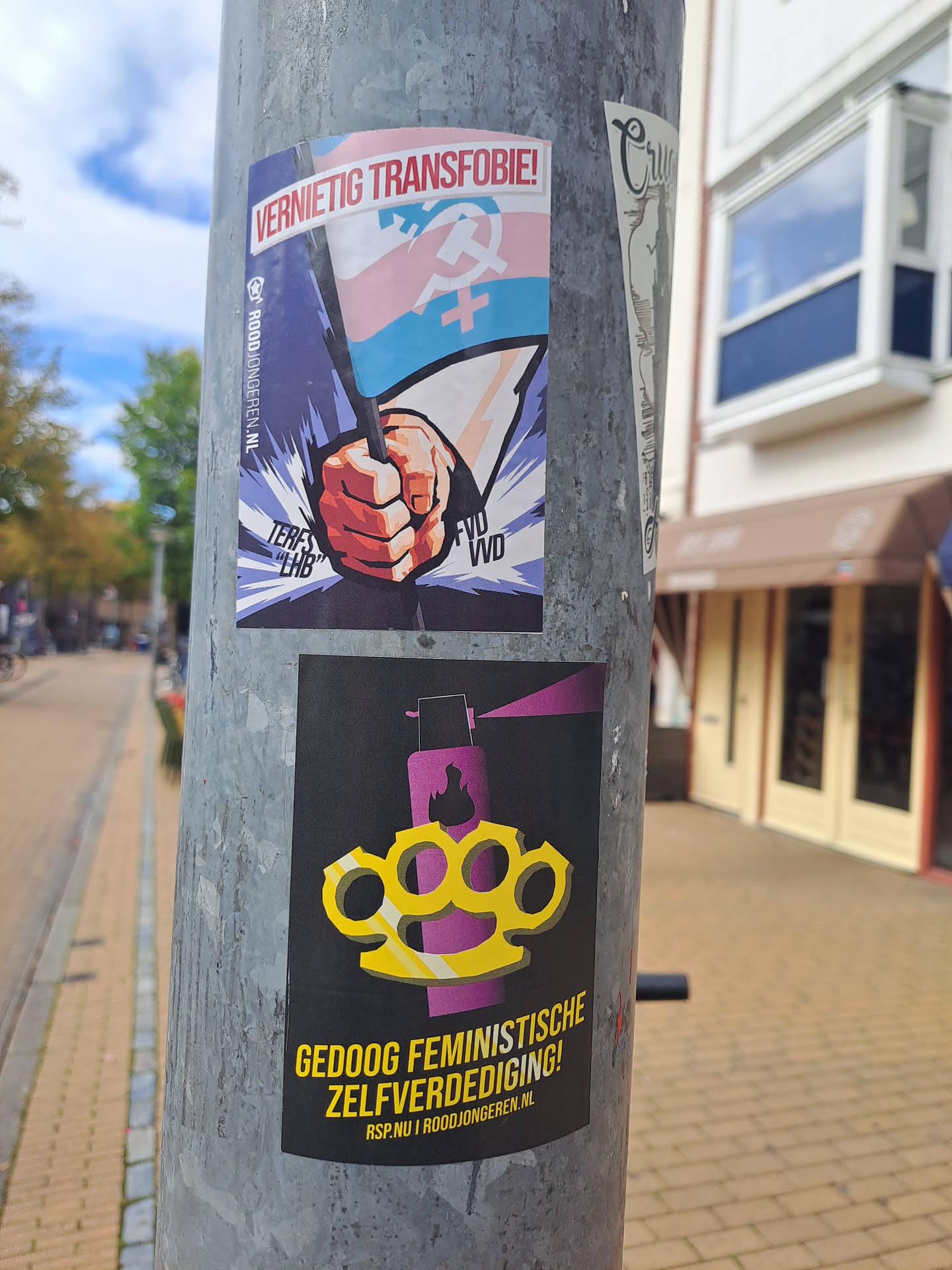 The central sticker is black with yellow and purple graphics. It features a pair of bright yellow brass knuckles superimposed over a stylized purple spray can with flames. Below the graphic, bold yellow text reads 'GEDOOG FEMINISTISCHE ZELFVERDEDIGING!' (Tolerate feminist self-defense!). At the bottom, smaller text indicates 'RSP.NU | ROODJONGEREN.NL'. Above this sticker, another sticker displays a hand holding a flag with the transgender colors (pink, white, light blue) and symbols of a hammer and sickle, and a Venus symbol. The text on this upper sticker reads 'VERNIETIG TRANSFOBIE!' (Destroy transphobia!).