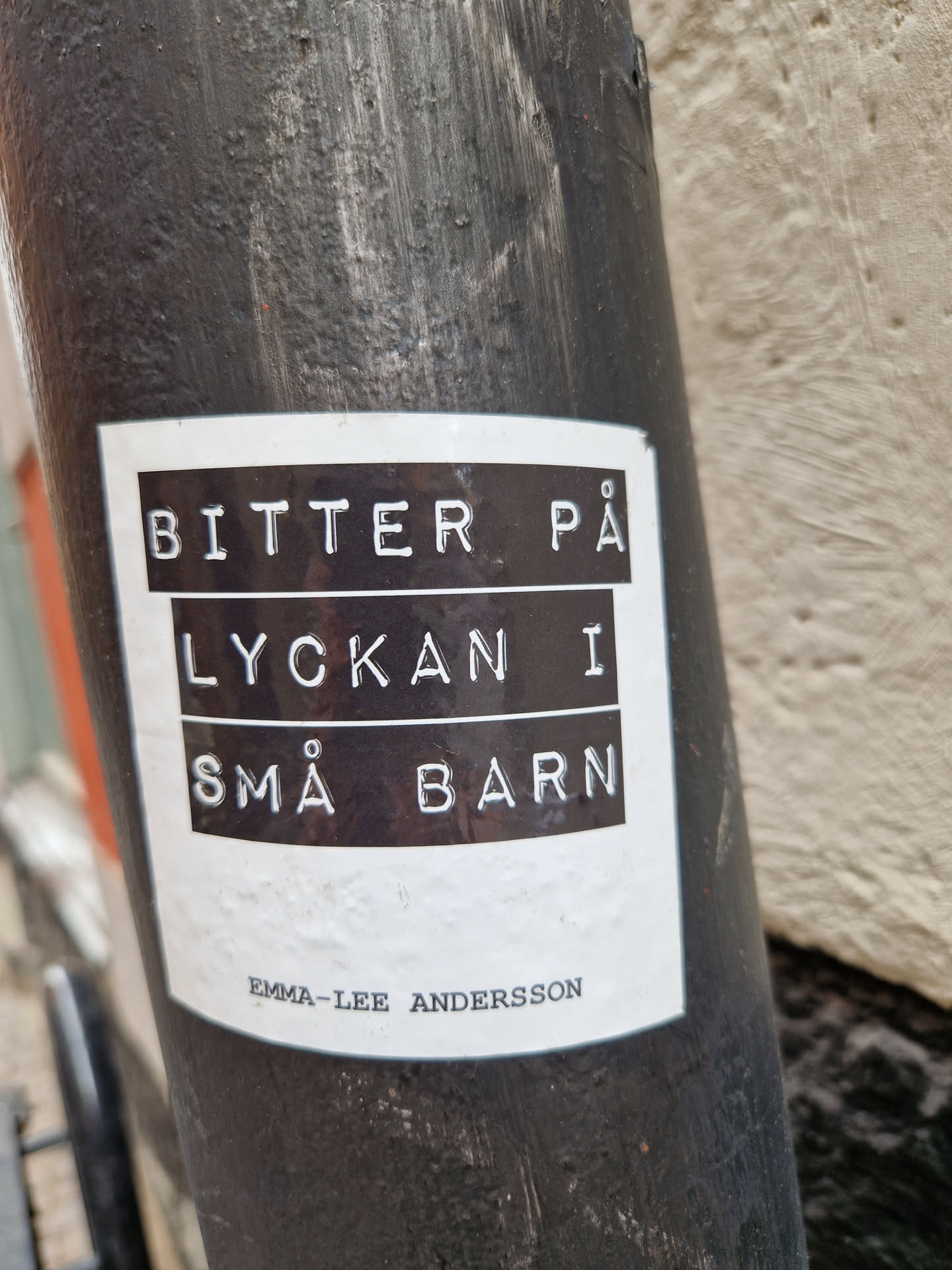 A black and white sticker with the text "Bitter på lyckan i små barn" (Swedish for "Bitter on happiness in small children") and the artist's name Emma-Lee Andersson.