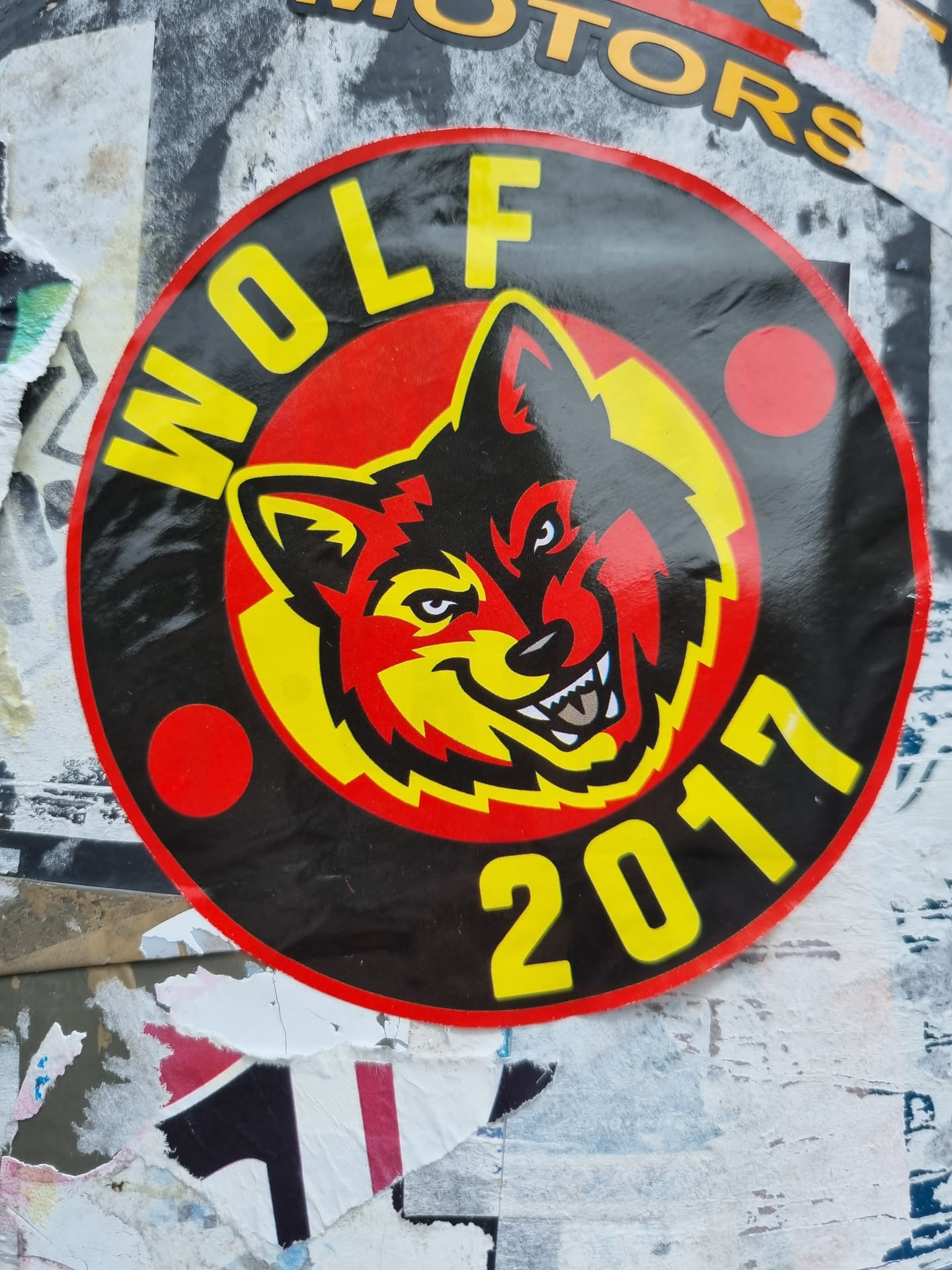 Close-up of a round sticker featuring a stylized wolf's head in red and yellow, set against a black background. The word "WOLF" is written in yellow uppercase letters at the top, and "2017" in yellow at the bottom. The sticker has a red border.