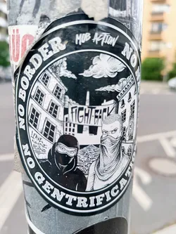 A black and white sticker with an illustration of two masked figures standing in front of buildings. The text around the circle says "No Border No Gentrification" with the words Mob Action at the top.  The words "Fight Back" are visible on a banner in the illustration. There is also smaller text that says "Leave no one behind".