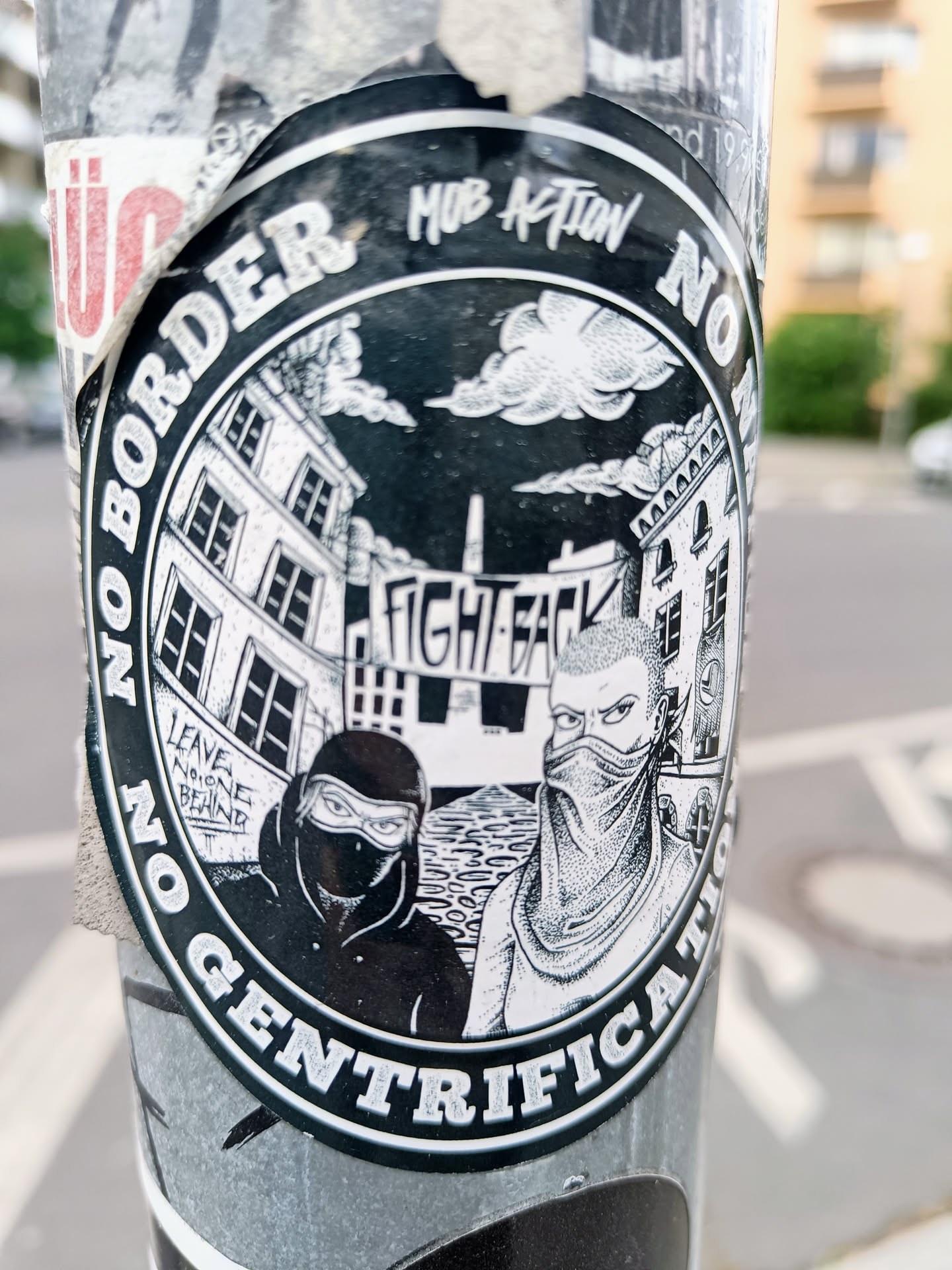 A black and white sticker with an illustration of two masked figures standing in front of buildings. The text around the circle says "No Border No Gentrification" with the words Mob Action at the top.  The words "Fight Back" are visible on a banner in the illustration. There is also smaller text that says "Leave no one behind".