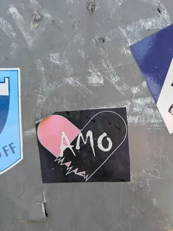 A black sticker featuring a stylized broken heart design. The upper left portion of the heart is filled with a light pink color, while the rest of the heart is outlined in white on a black background. The word "AMO" is written in white, stylized letters across the center of the heart. The bottom of the heart is jagged, resembling a broken edge.