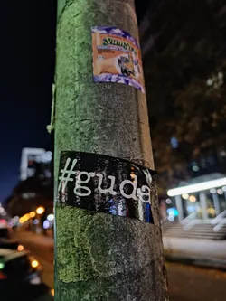 A black and white sticker with the text #guda is affixed to a lamppost at night. The sticker has a slightly reflective surface and is partially damaged.