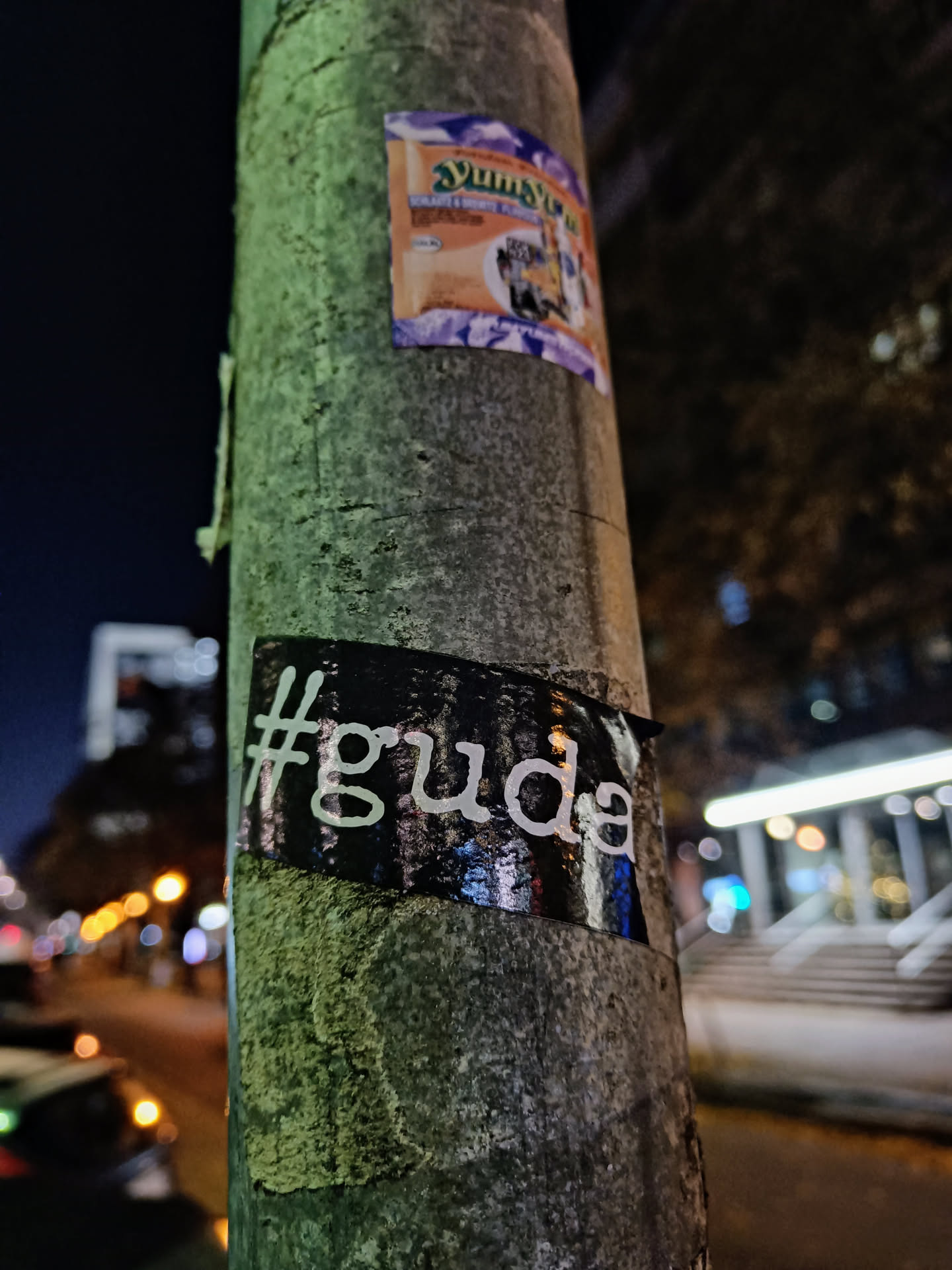 A black and white sticker with the text #guda is affixed to a lamppost at night. The sticker has a slightly reflective surface and is partially damaged.