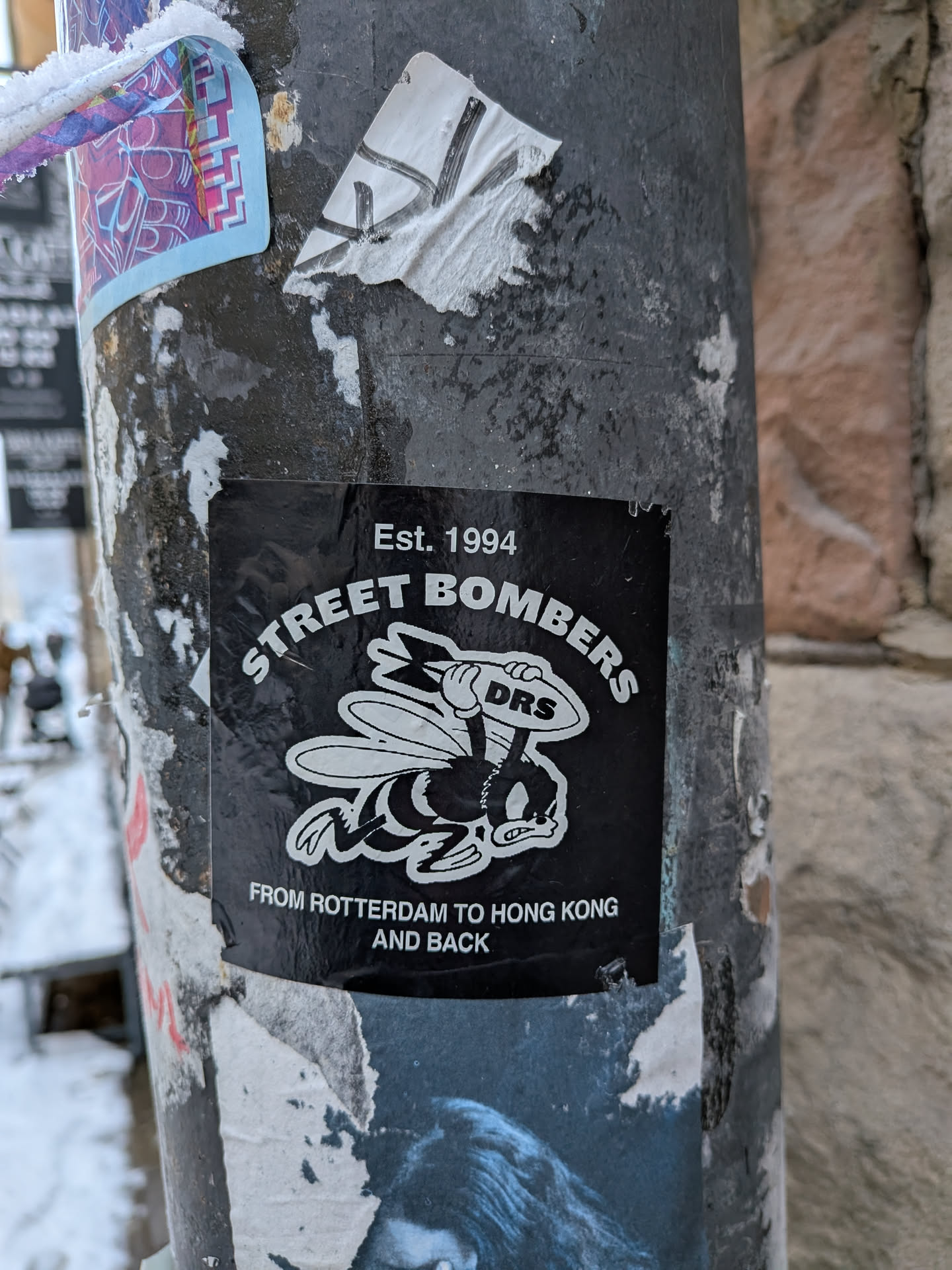 A square black sticker featuring a white cartoonish bee with an angry expression, holding a bomb labeled 'DRS'. The text 'Est. 1994' is at the top, 'STREET BOMBERS' arcs above the bee, and 'FROM ROTTERDAM TO HONG KONG AND BACK' is at the bottom, all in white.