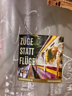 Street sticker A square sticker featuring a photograph of a young woman leaning out of a train window. The main text on the sticker says "Züge statt Flüge" which translates from German to "Trains instead of planes".  A small penguin logo is visible at the bottom. A website and Instagram handle are also included. 