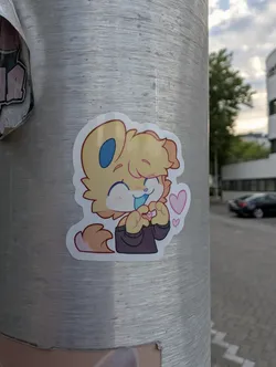 A die-cut sticker of a chibi-style yellow furry character with blue inner ears and tongue. The character wears a dark sweater, smiles with closed eyes, and forms a heart shape with its paws, flanked by small pink hearts.