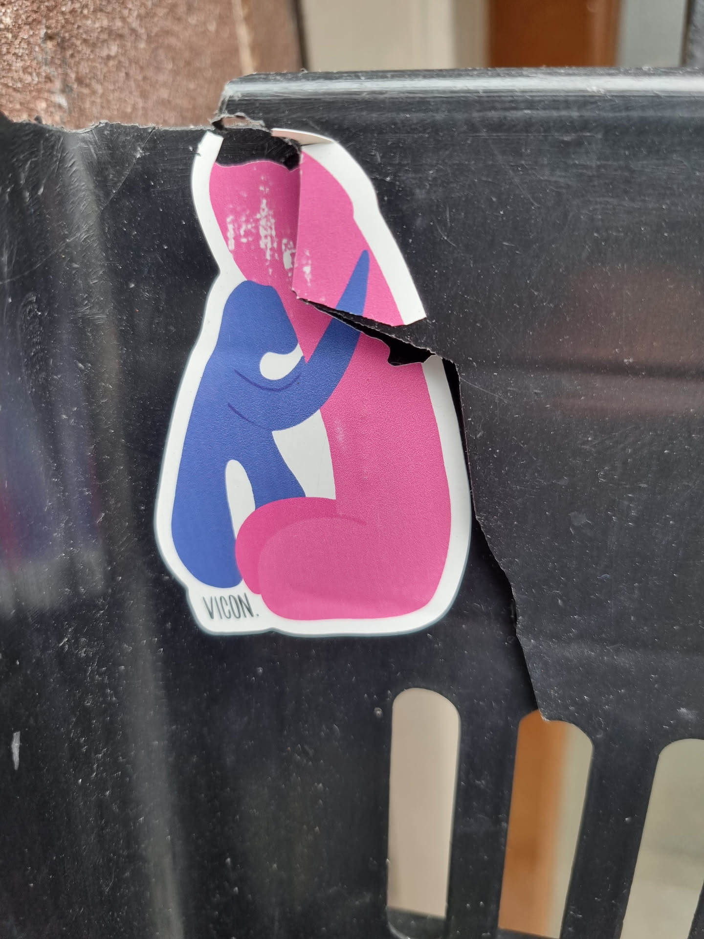 This sticker depicts a simple, stylized illustration of two figures, one blue and one pink, in a tender embrace. The style is minimalist and modern. The sticker itself is damaged, with a significant tear along the right side, revealing the surface beneath. Despite the damage, the central image remains largely intact.  A small text, 'VICON.', appears at the bottom of the design.