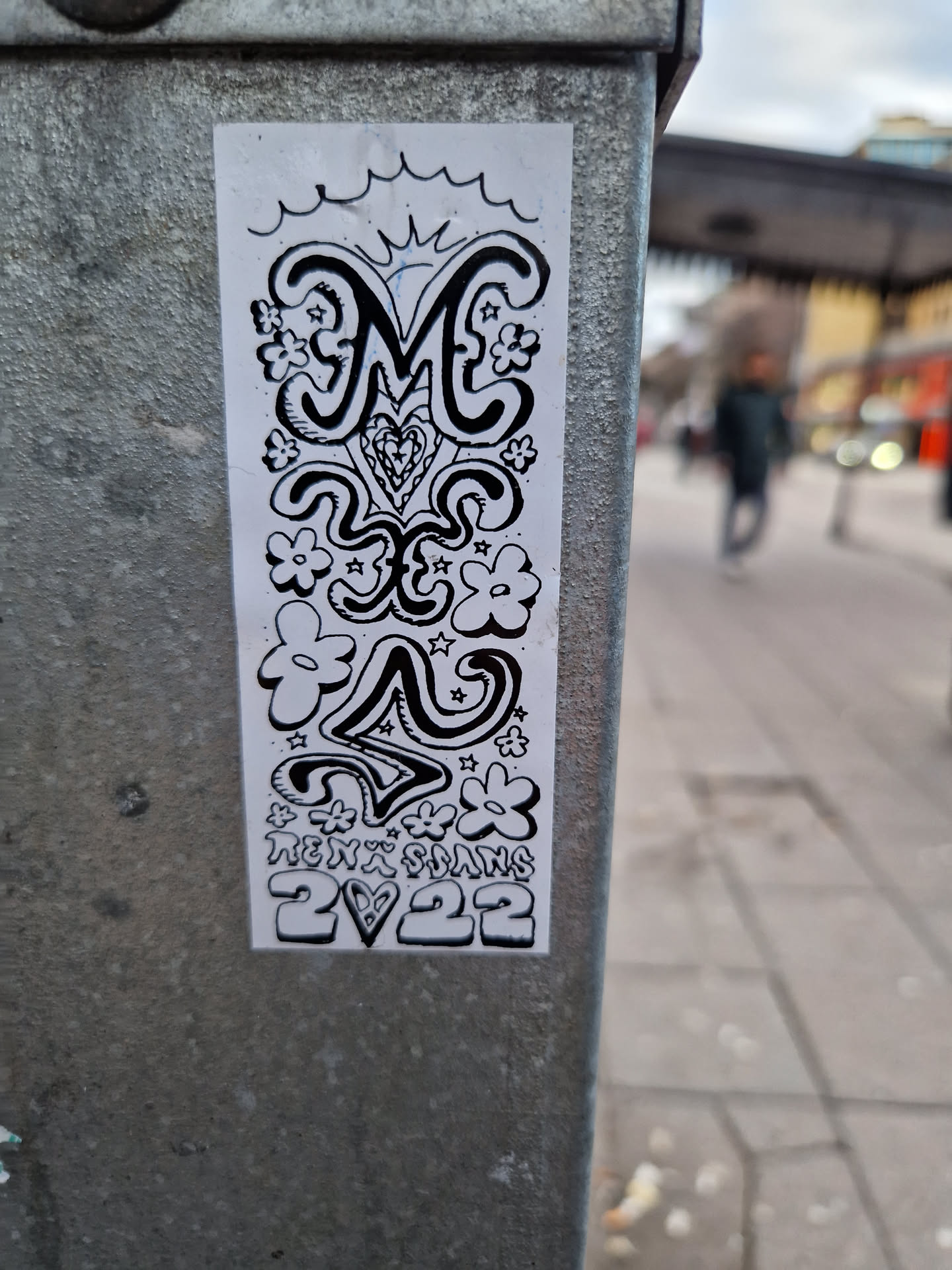 This sticker is a black and white drawing, seemingly hand-drawn, with a central focus on a stylized letter 'M'.  It features a whimsical design with flowers, stars, and abstract shapes surrounding the letter. The artist's name and year are included at the base.