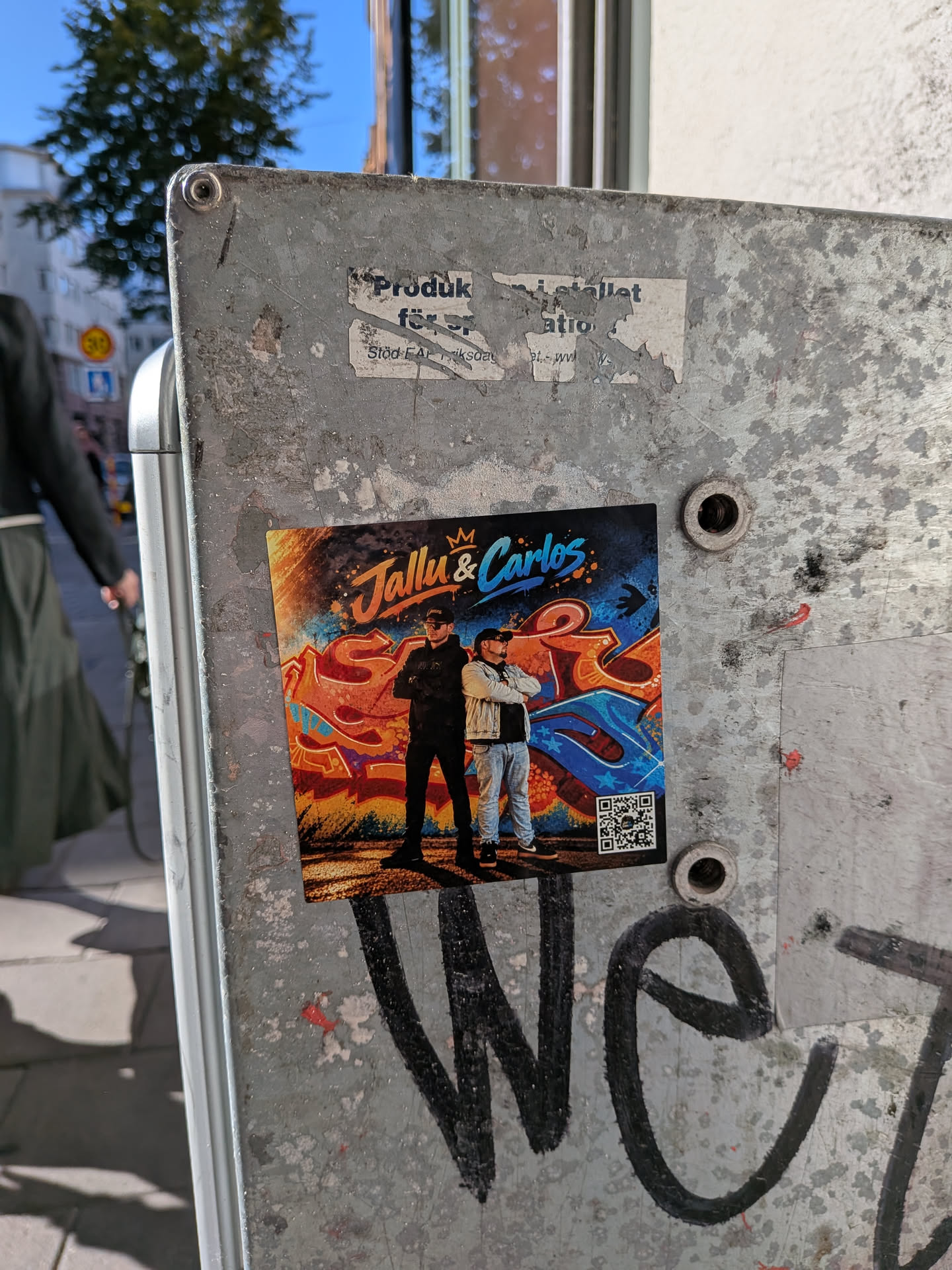 A square color sticker showing two men standing back-to-back in front of a vibrant graffiti wall with bold orange, blue, and red tones. The man on the left is dressed in black streetwear, while the man on the right wears a denim jacket and light blue jeans. Above them, the text 'Jallu & Carlos' is written in stylized brush script beneath a small gold crown. A small QR code is visible in the lower right corner.