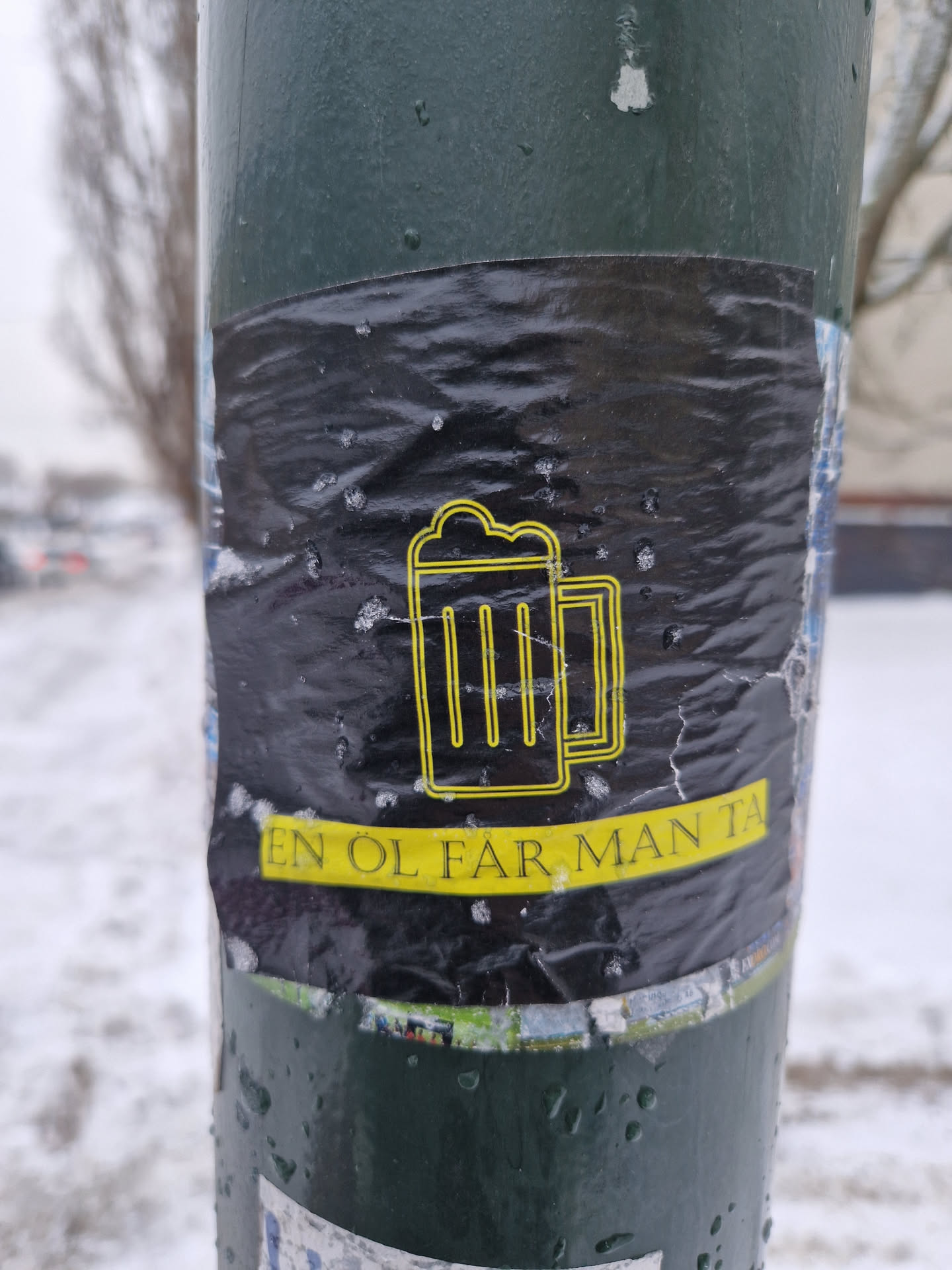 A black and yellow sticker featuring a beer mug graphic and the text "En öl får man ta!" which translates to "One beer you can take!" in Swedish. The sticker is affixed to a lamppost in a snowy setting.