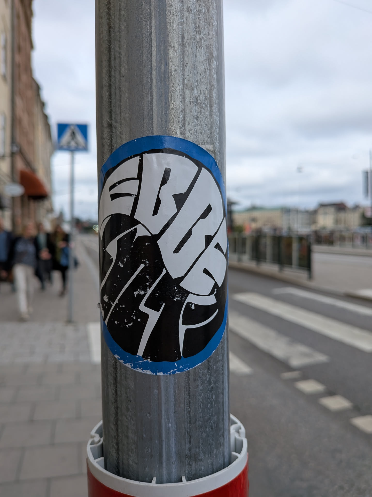The sticker is circular with a prominent blue border. Inside the border, the design features bold, stylized white lettering that appears to be a word or name, set against a black background. The white lettering has sharp angles and thick strokes. The black background has a slightly distressed texture with small white speckles, suggesting wear or intentional design. The sticker is affixed to a textured, weathered metal pole.