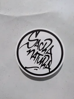 A circular sticker features a stylized black and white design. The words "Sack Natura" are rendered in a unique, almost graffiti-like font, positioned within the circle. The sticker is placed on a neutral, slightly textured surface.