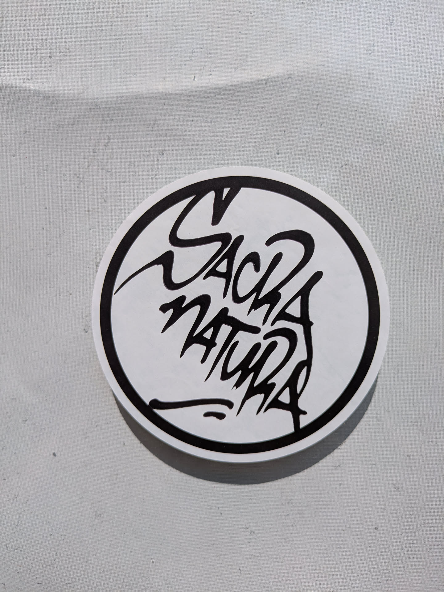 A circular sticker features a stylized black and white design. The words "Sack Natura" are rendered in a unique, almost graffiti-like font, positioned within the circle. The sticker is placed on a neutral, slightly textured surface.