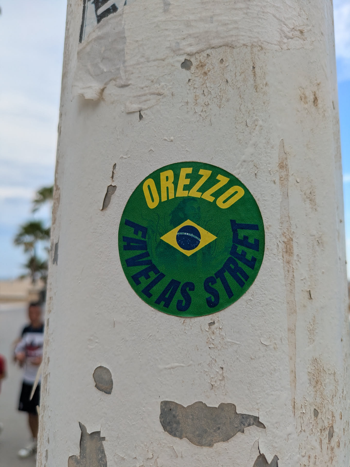 The sticker is on a light pole. It is round and colored green with yellow text that reads "OREZZO" at the top and "FAVELAS STREET" at the bottom. In the center is the Brazilian flag. The pole itself is white, with patches of peeling paint revealing a darker surface underneath.