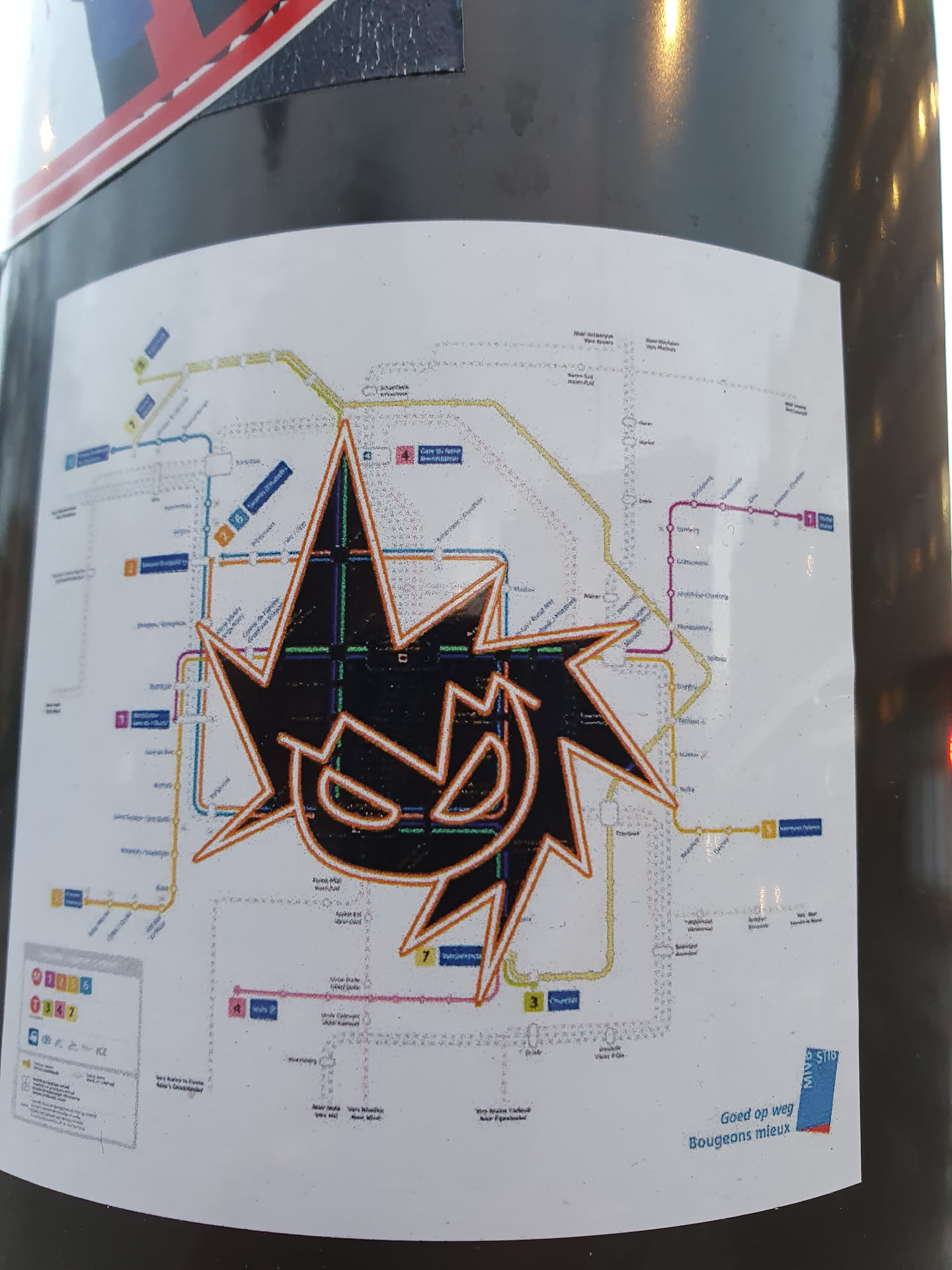 A black and orange stylized graphic sticker is placed over a public transport map. The sticker features a jagged, starburst-like design with a face-like element in the center. The map appears to be of a tram or train system, with various lines and station names visible. The bottom right of the map features text in Dutch indicating the location, which seems to be Bougeniers plein.