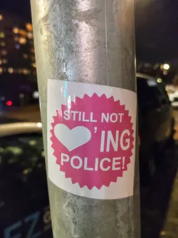 A pink sticker with a white heart and the text "Still not loving police!" on a metal pole.