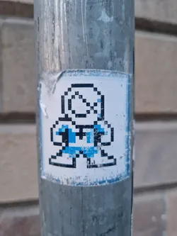 A sticker featuring a pixelated character in blue and black on a white background. The sticker is attached to a gray metal pole and shows some wear.
