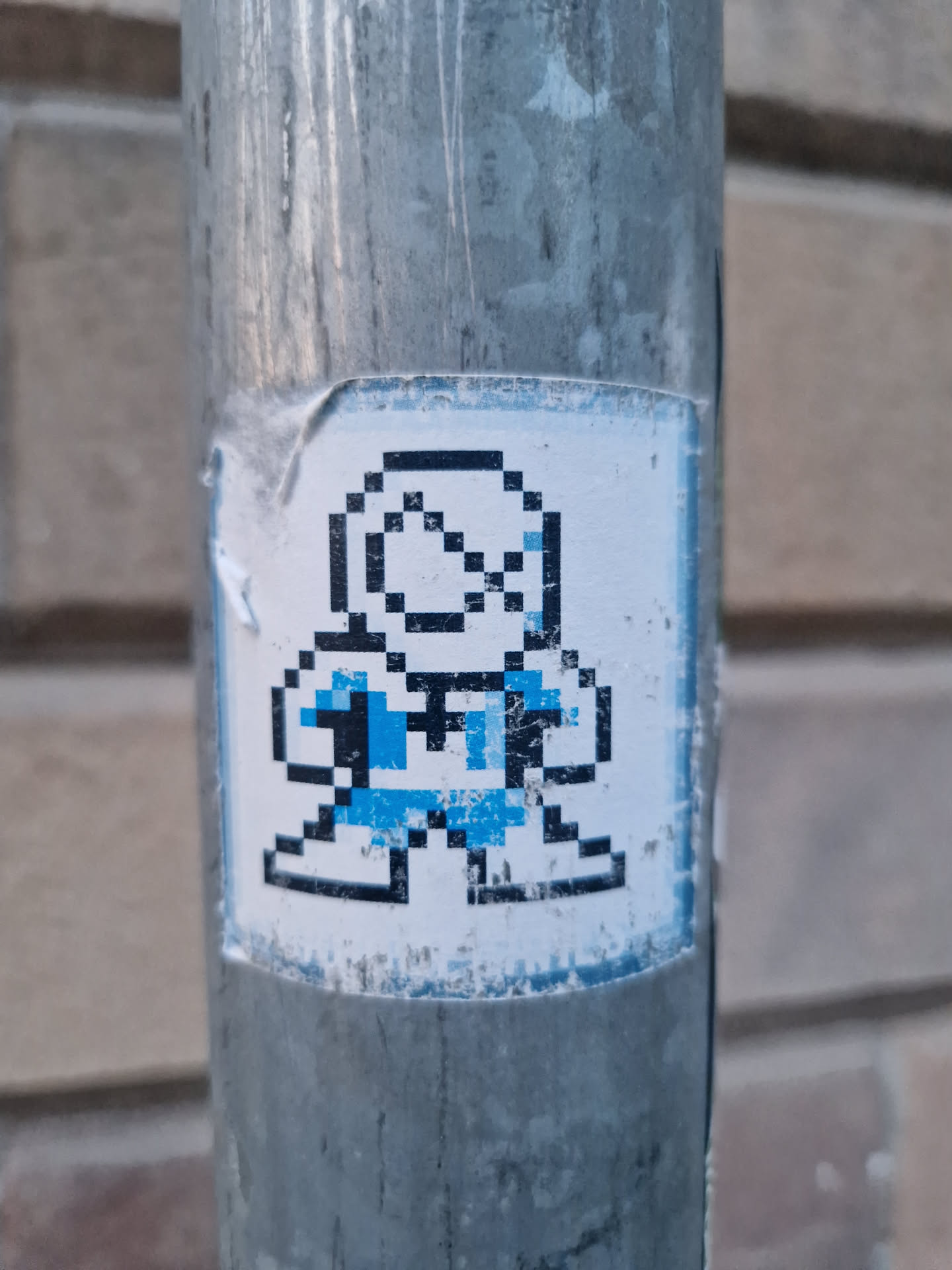 A sticker featuring a pixelated character in blue and black on a white background. The sticker is attached to a gray metal pole and shows some wear.