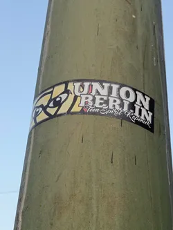 A sticker featuring the text \"UNION BERLIN\" with \"Teen Spirit Köpenick\" written below it in a smaller font. The sticker has a graphic element to the left of the text. It's affixed to a weathered green pole.