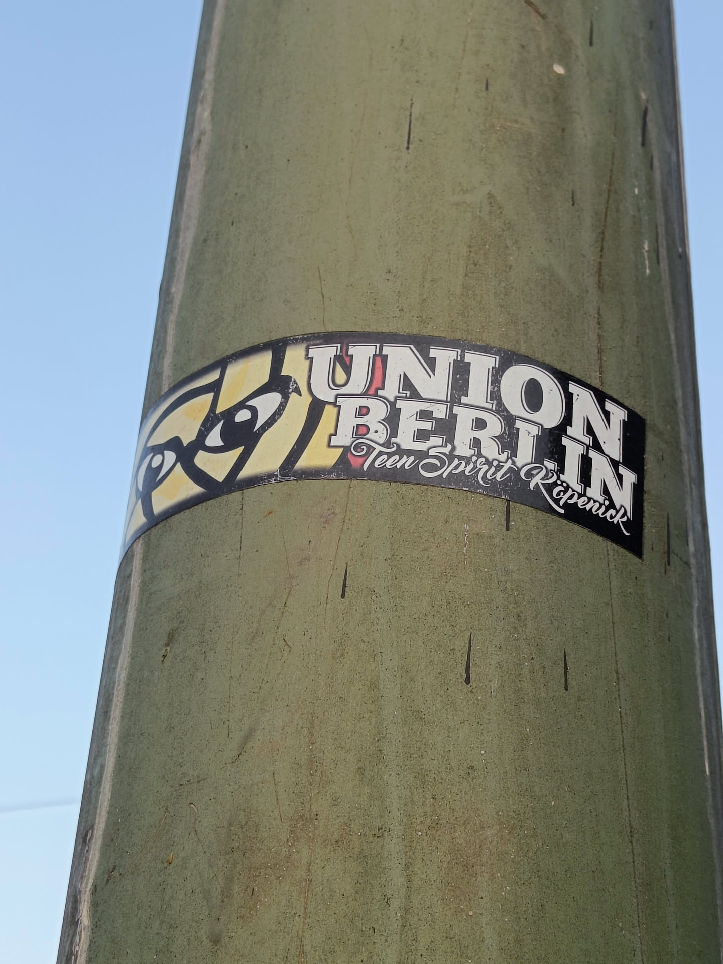 A sticker featuring the text \"UNION BERLIN\" with \"Teen Spirit Köpenick\" written below it in a smaller font. The sticker has a graphic element to the left of the text. It's affixed to a weathered green pole.