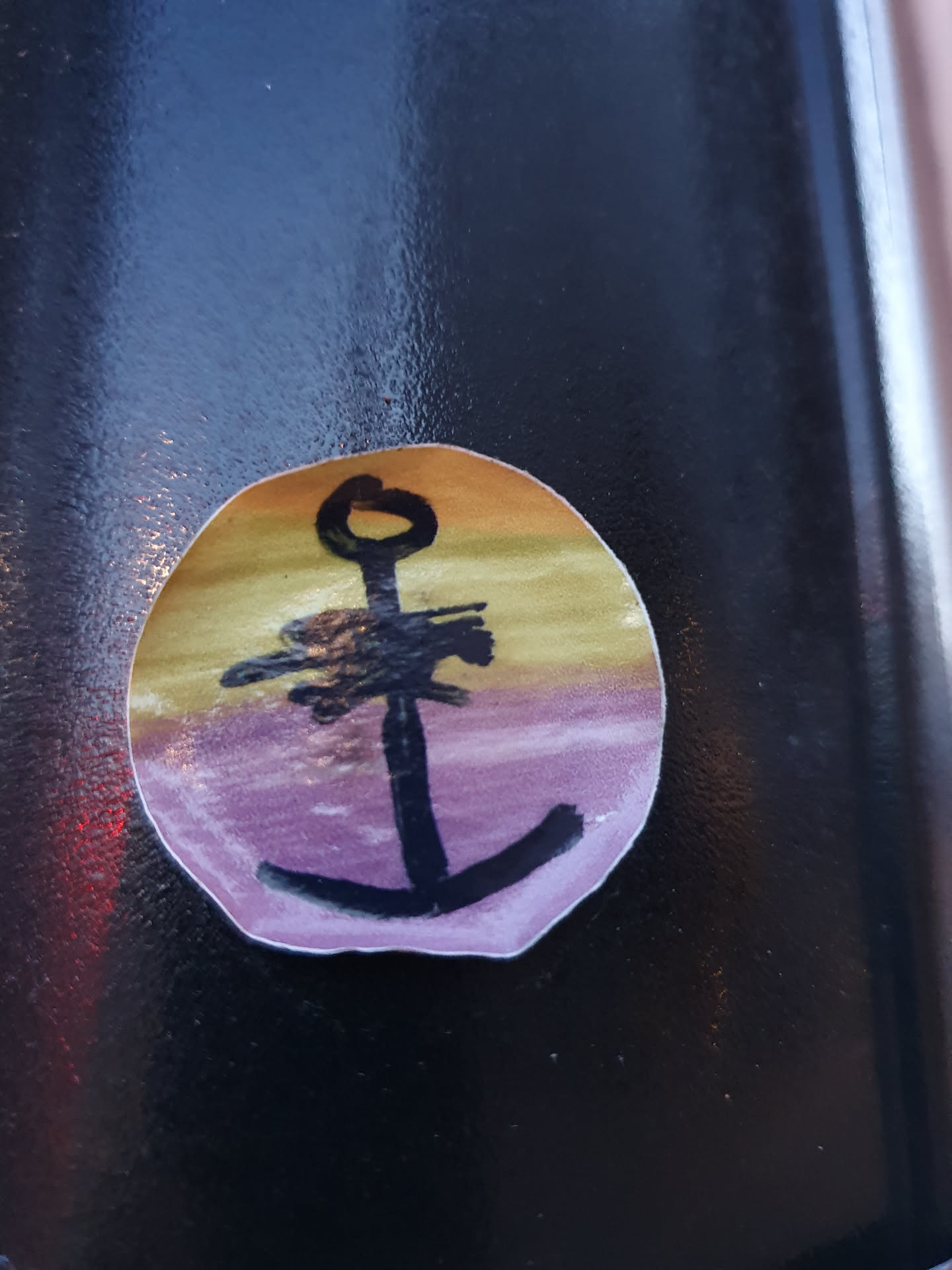 A small, round sticker featuring a simple drawing of an anchor. The anchor is black, and the background is a gradient of yellow, orange, and purple, reminiscent of a sunset.  The style is child-like or naive.