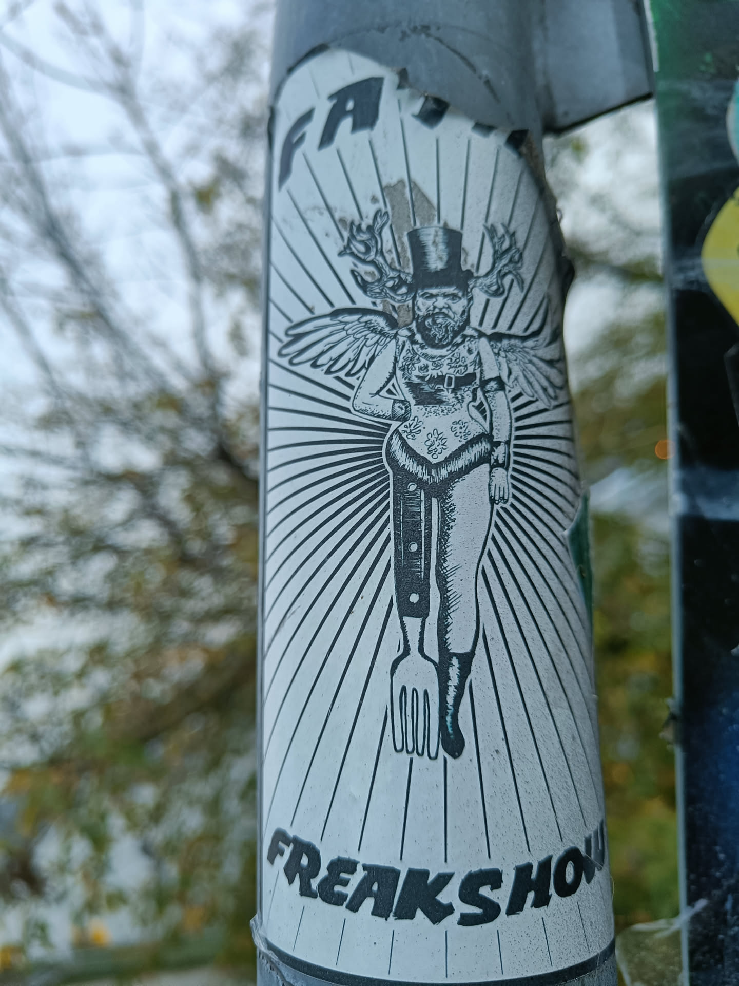 A white sticker with black ink illustration of a figure with antlers growing out of its head, wearing a top hat and wings. The figure has legs ending in a fork and knife. The sticker has the word "FREAKSHOW" at the bottom. 