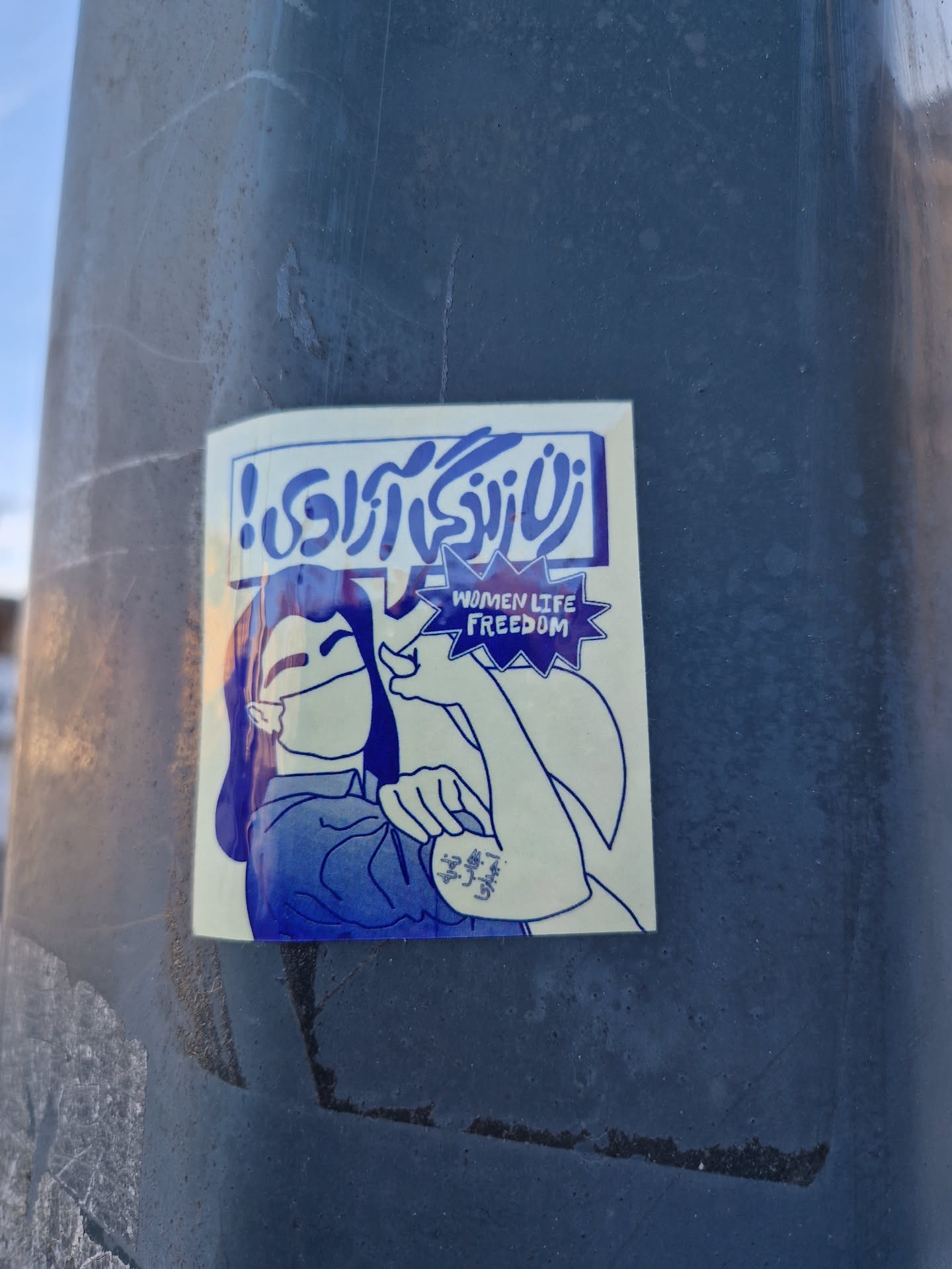 A blue and white sticker featuring a woman in a Rosie the Riveter pose.  The sticker includes Farsi text and the words "Women Life Freedom".