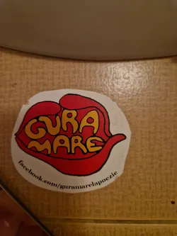 Round sticker with an illustration of a mouth saying Gura Mare. The text is in yellow and red, and the mouth is red. There is a Facebook link at the bottom.