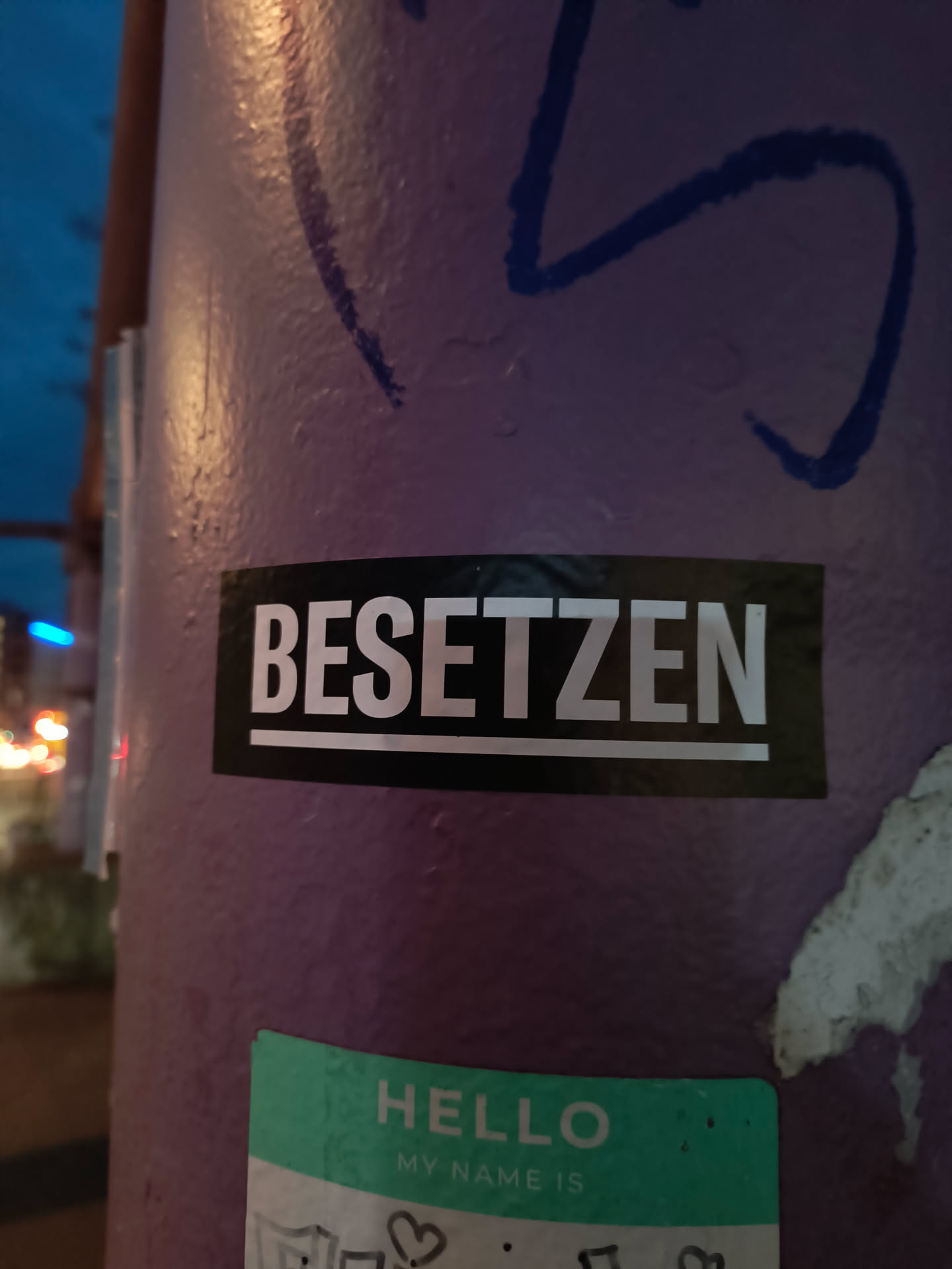 The sticker is rectangular with a black background and white, sans-serif text. The word 'BESETZEN' is centered on the sticker, underlined by a thin white line. The text is large and blocky, giving it a strong, impactful appearance. The sticker appears to be adhered to a purple, textured surface, possibly a pole, which also has some blue graffiti on it.